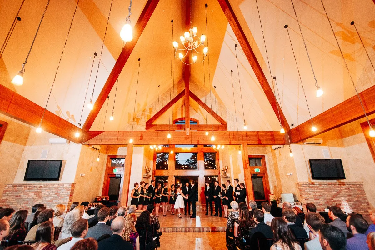 A wedding ceremony in a warmly lit, triangular-roofed hall. Couples are at the altar, surrounded by bridesmaids and groomsmen. Guests fill the seats in a celebratory atmosphere.