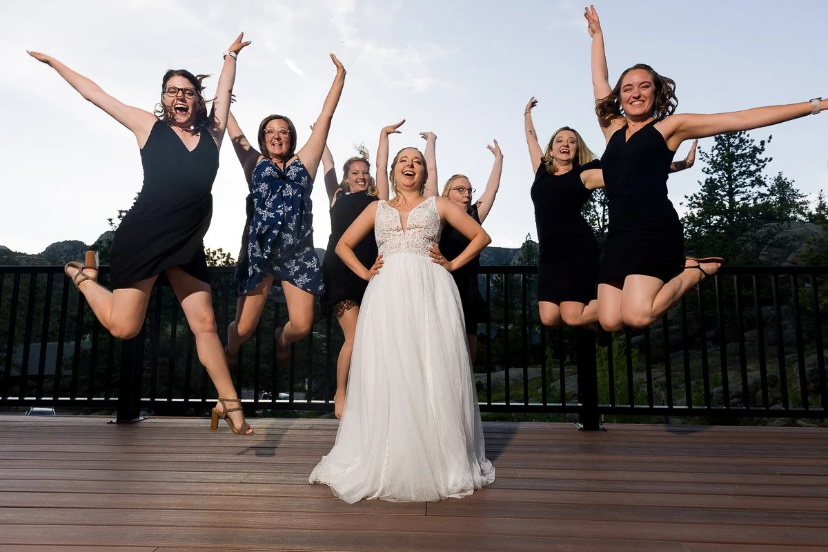 A joyful bride in a white gown stands on a deck, surrounded by six bridesmaids in black dresses, all jumping mid-air, with trees in the background.