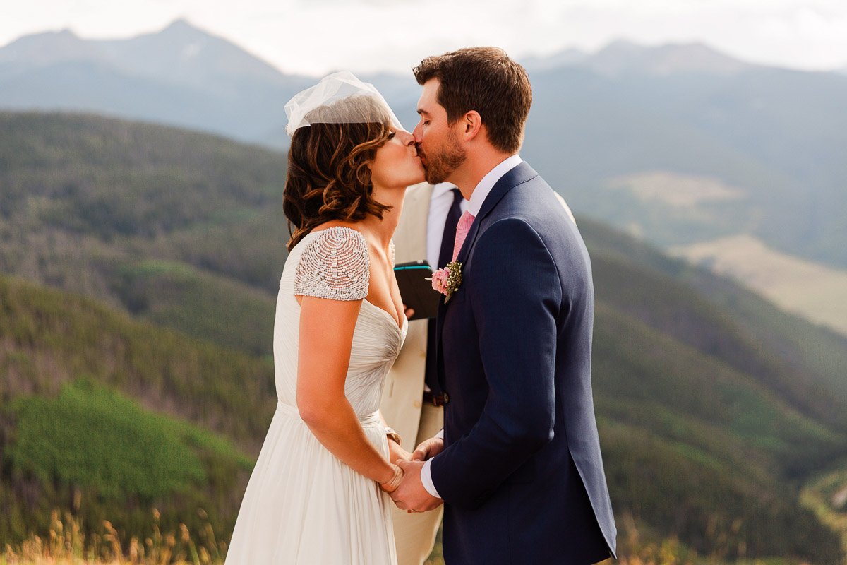 A bride in a white gown and veil kisses a groom in a navy suit and pink tie, set against a mountainous backdrop. They hold hands, conveying joy.