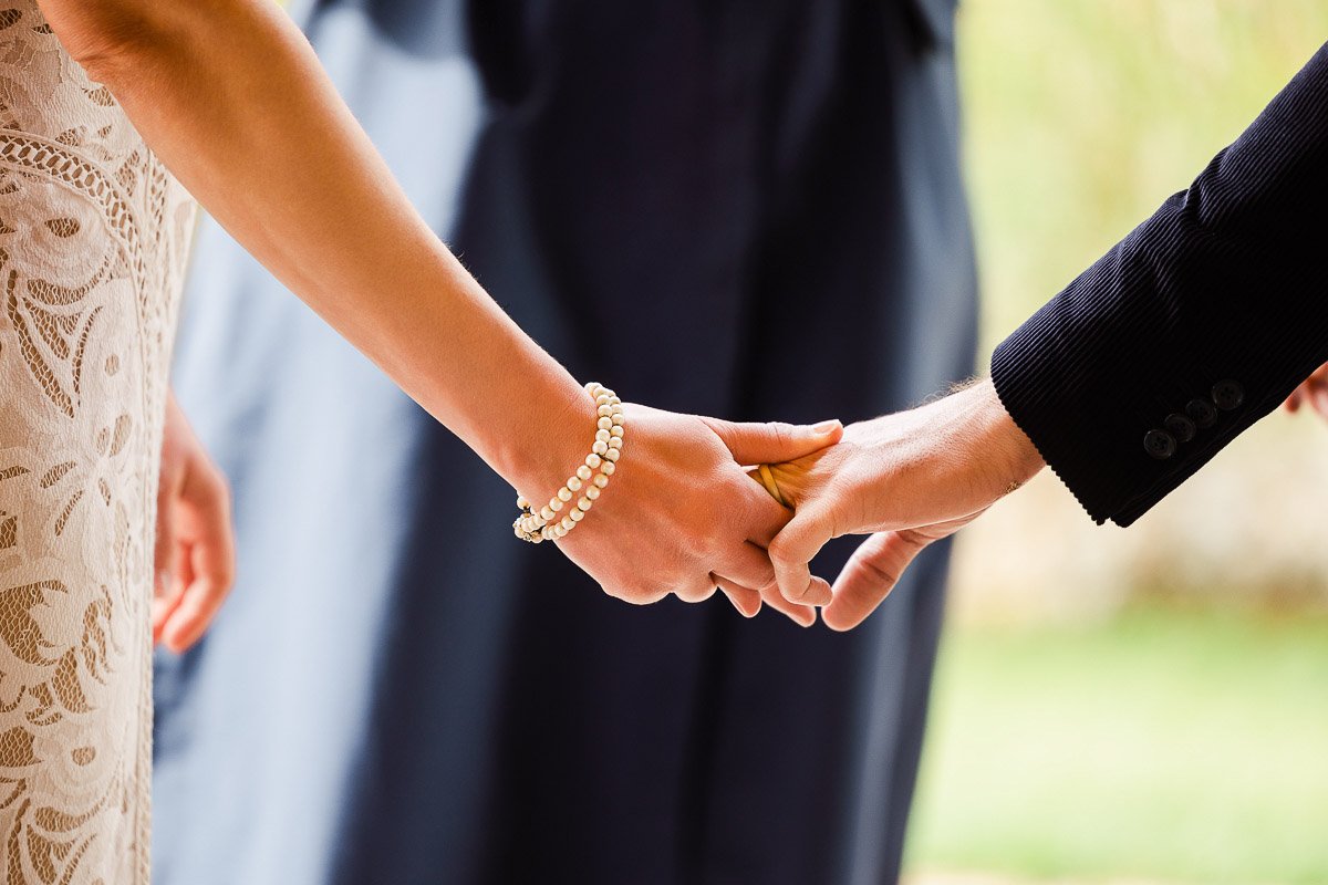 Close-up of two people holding hands during a ceremony. One wears a lace dress and pearl bracelet, the other's in a dark suit, conveying love and commitment.
