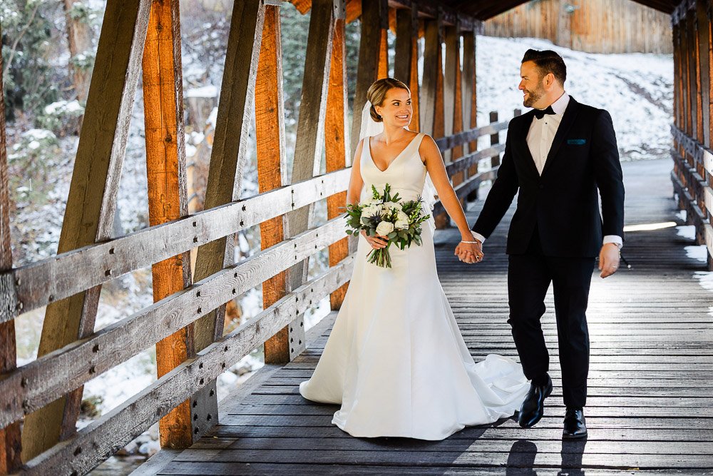A newlywed couple joyfully walks hand in hand on a rustic wooden bridge. The bride wears a white gown and holds a bouquet; the groom is in a black suit.