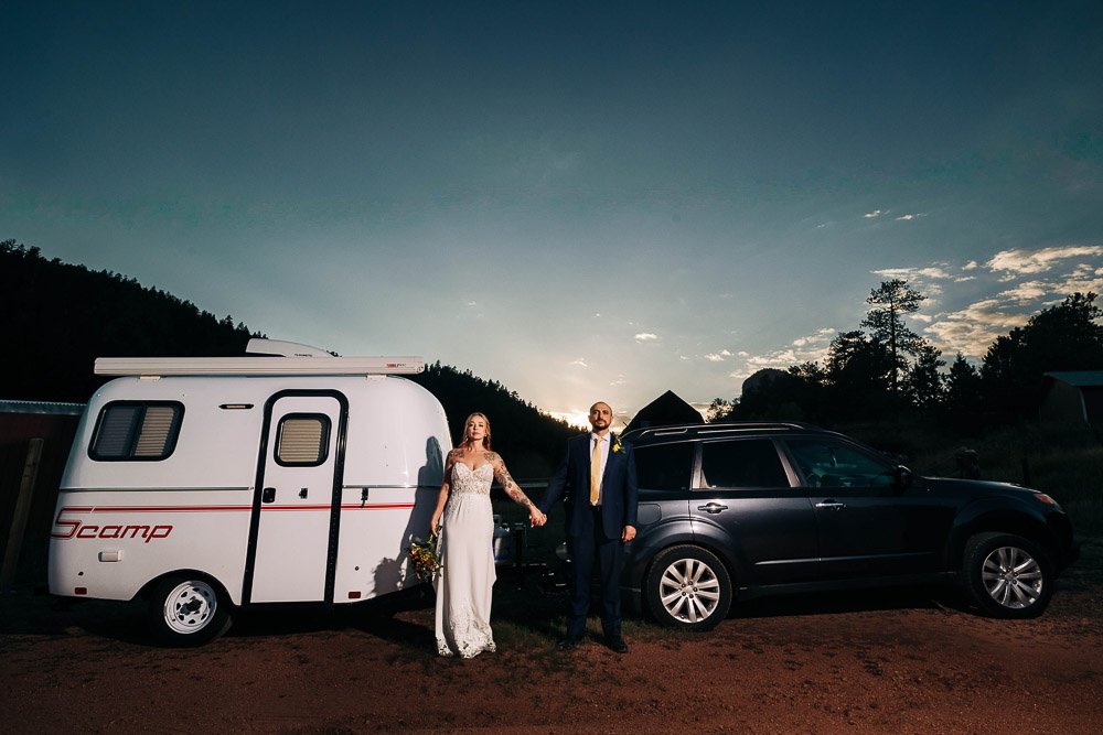 Bride and groom holding hands, standing beside a compact white trailer and dark SUV at dusk. The scene conveys adventure and romance.