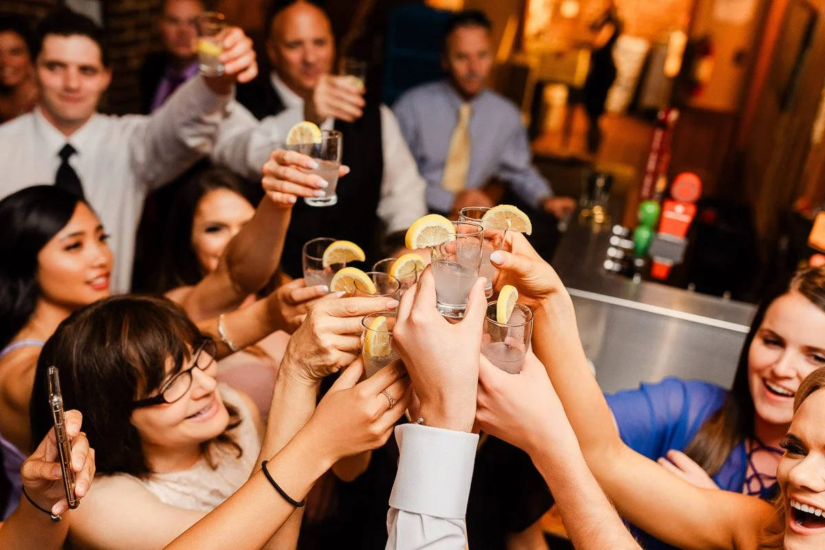 A group of people toasting with lemon-adorned cocktails in a lively bar setting, exuding joy and celebration. Mixed ages, varied attire.