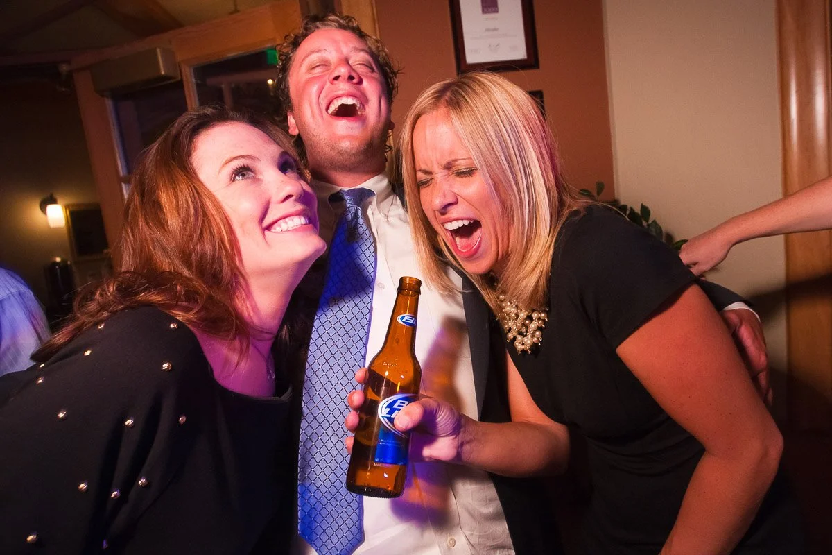 A joyful group of three friends laughing at a party. A woman in black holds a beer, while others lean in close, exuding a lively, cheerful atmosphere.