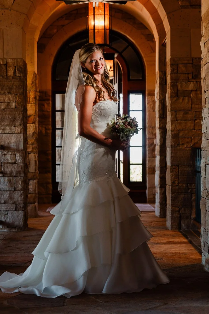 Bride in a white dress stands in a stone hallway, holding a bouquet. Soft lighting creates a warm, romantic atmosphere. Smiling expression during a Sanctuary Golf Course wedding in Sedalia, Colorado.