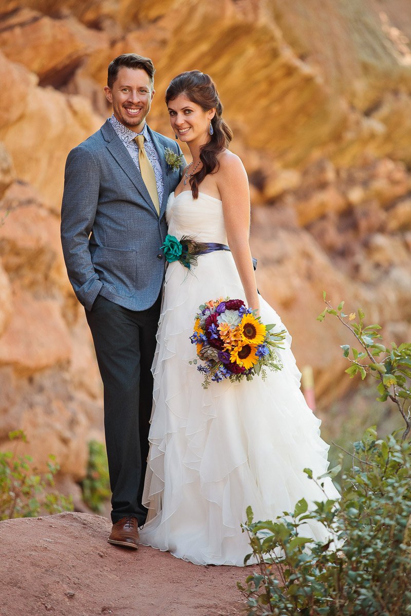 Bride in a white gown with a blue sash stands beside a groom in a blue suit, holding hands in a scenic canyon setting, exuding romance.