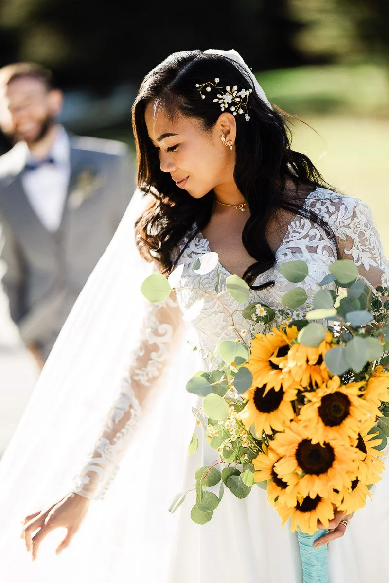 Bride in a lace gown holding sunflowers, smiling gently. Her hair is adorned with a floral clip. A blurred groom in a suit stands in the background.