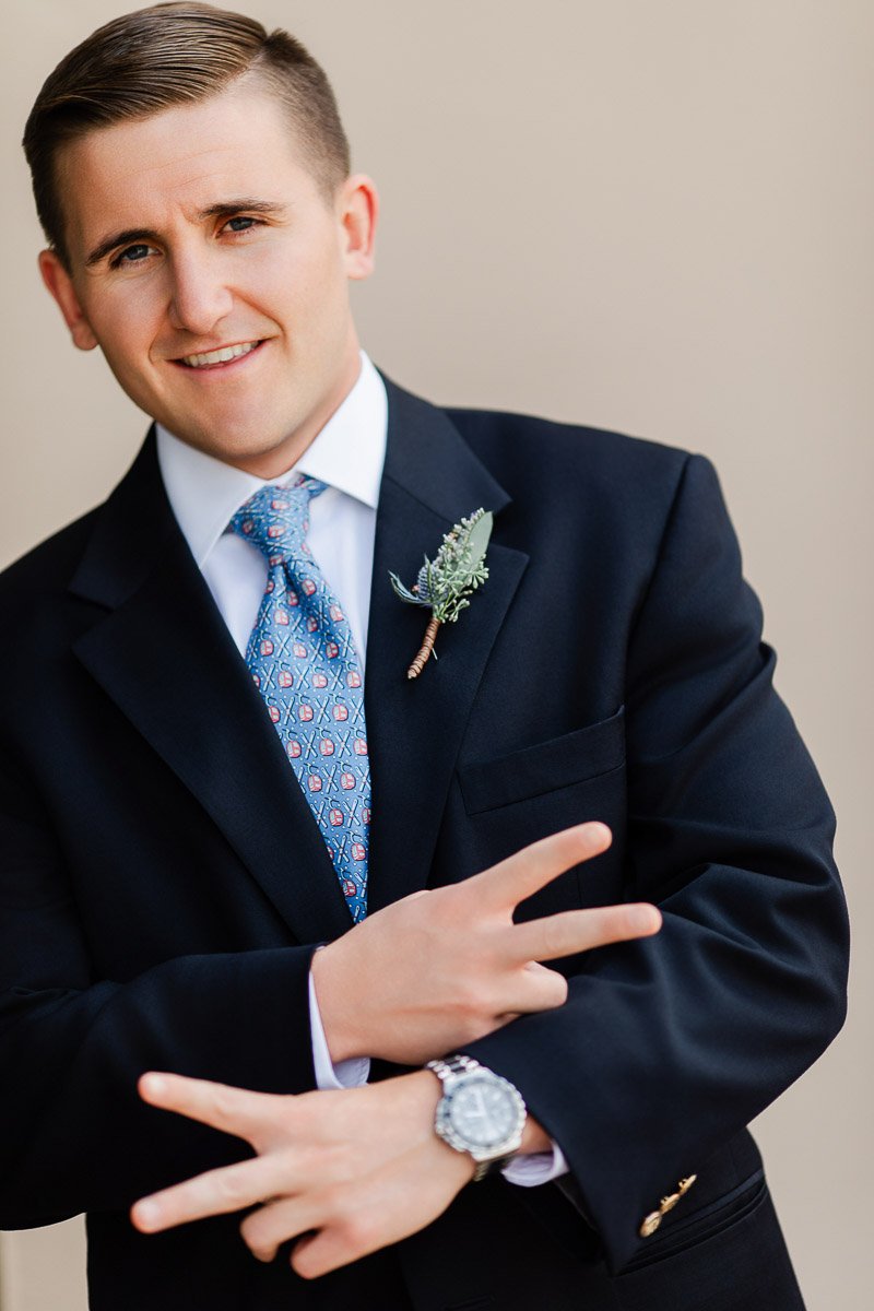A smiling man in a black suit and patterned blue tie strikes a playful pose with crossed arms. He has a small boutonniere on his lapel, conveying a cheerful mood.