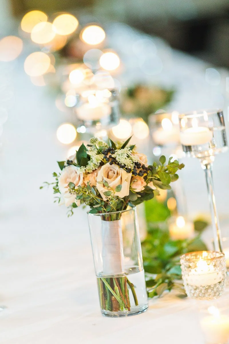 A table with elegant floral centerpieces featuring pale roses and greenery in clear vases, surrounded by softly glowing candles, creating a romantic atmosphere.