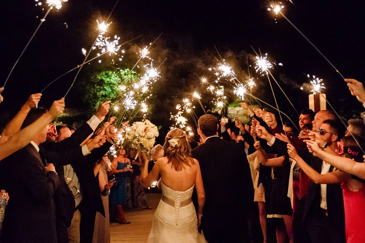 A bride and groom walk through a night-time aisle of guests holding sparklers. The scene is festive and joyful, creating a celebratory atmosphere.