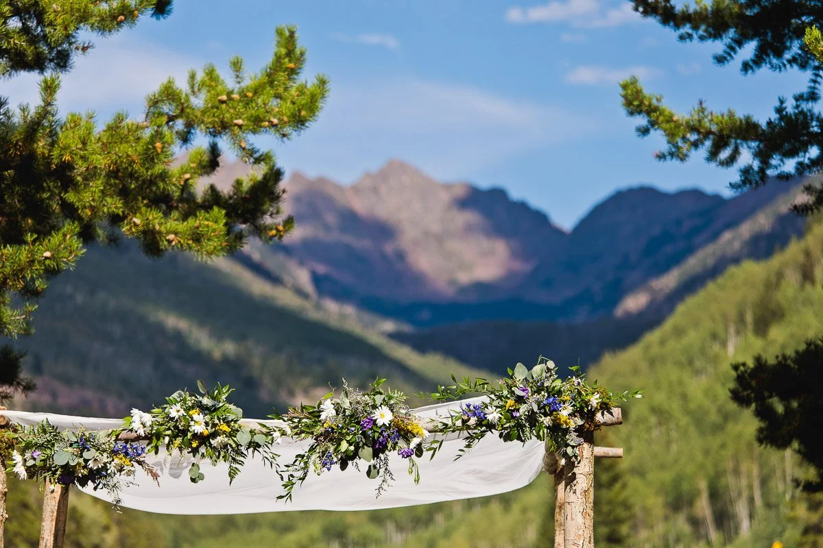 A wooden arch adorned with white fabric and lush greenery sits under a clear blue sky, framed by pine trees and mountains, evoking tranquility and natural beauty.