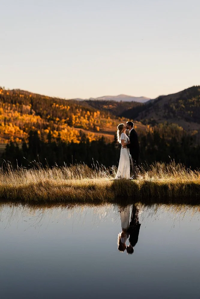 A couple embracing near a calm lake at sunset, with their reflection visible. Autumn trees and rolling hills create a warm, romantic backdrop.