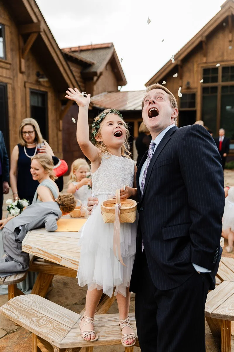 A joyful young girl in a white dress and floral crown, throws flower petals into the air. A man in a suit stands beside her, smiling. Celebration.