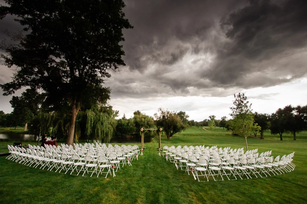 Outdoor wedding setup with two semi-circles of white chairs on green grass under dark, stormy clouds. An archway stands between the chairs.