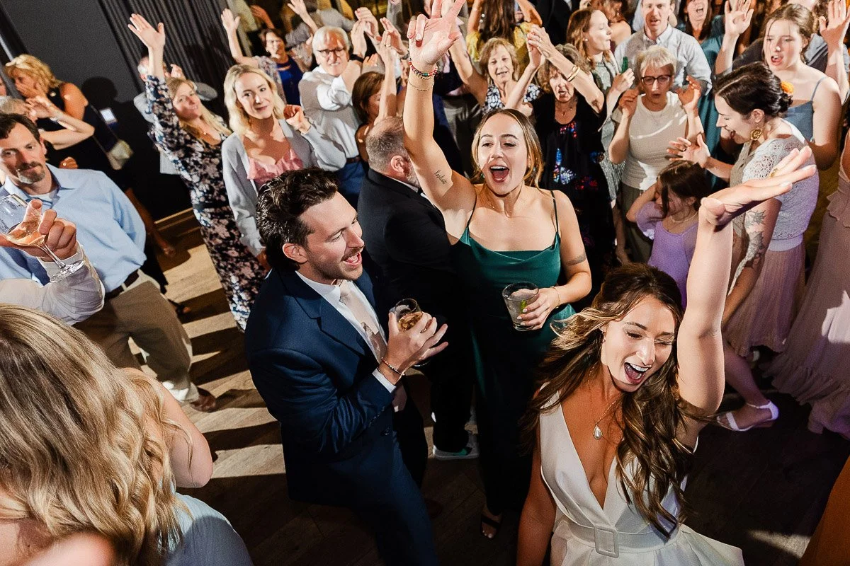 A joyful crowd dances at a wedding reception. People, dressed in formal attire, raise their hands, smiling and cheering in a lively, festive atmosphere.