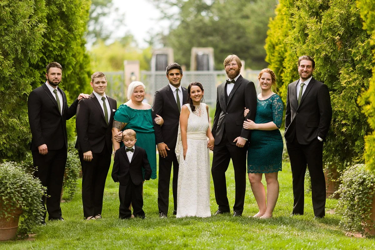 A wedding party stands in a lush garden, with the bride in a white dress, the groom and others in suits, and greenery surrounding them, smiling warmly.
