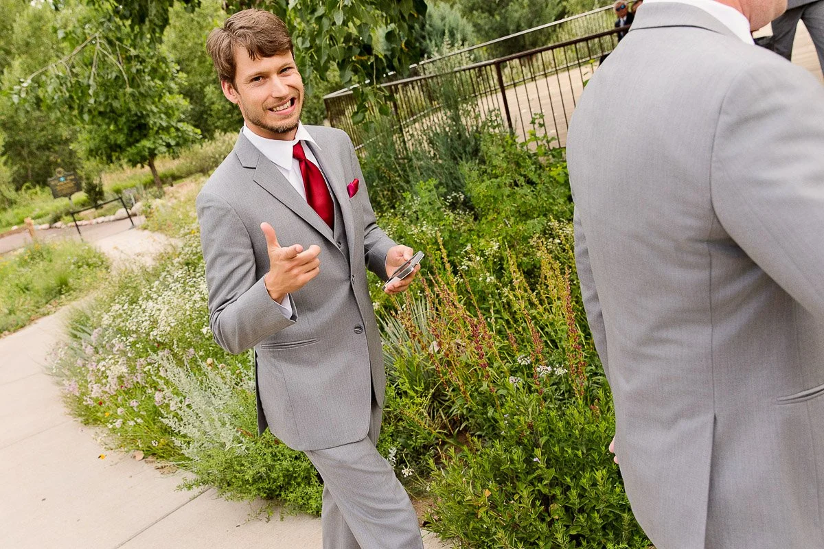 A man in a gray suit with a red tie smiles and gestures playfully while holding his phone. He stands on a path bordered by lush greenery.