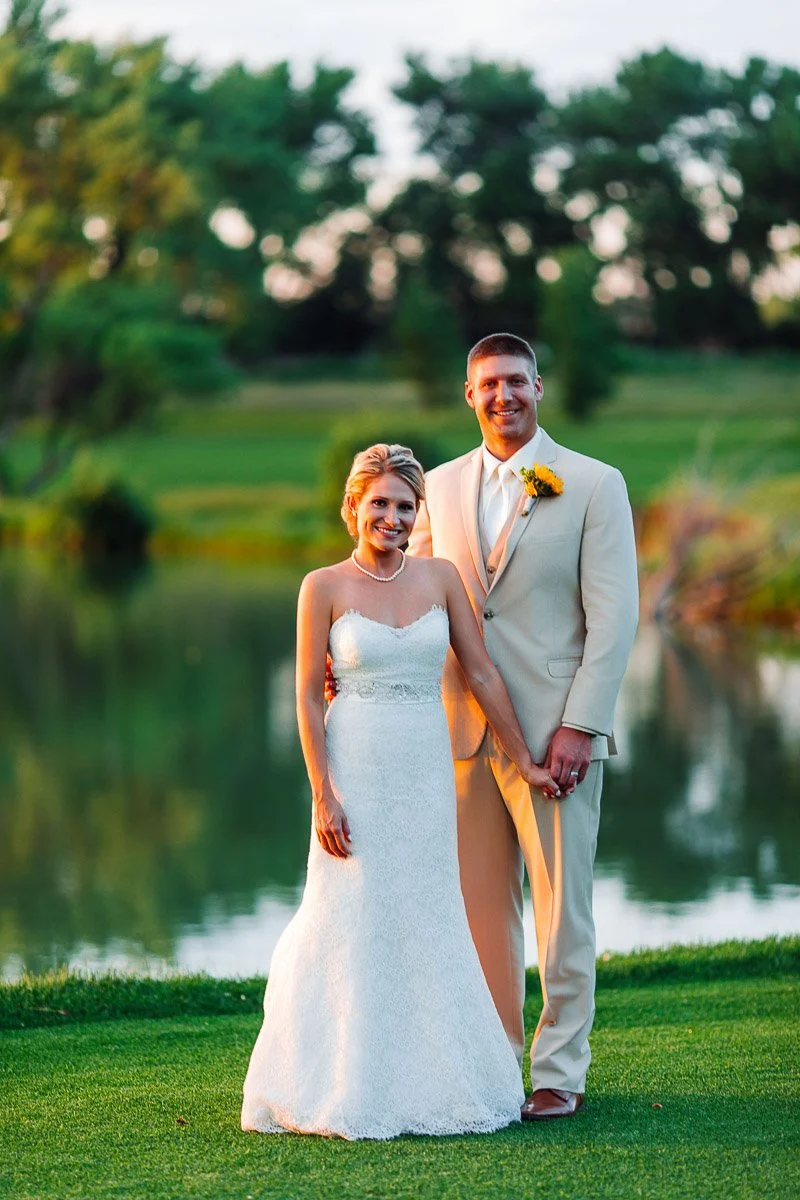A smiling bride and groom stand hand-in-hand on lush grass by a serene lake, with trees in the background. The setting is peaceful and romantic.