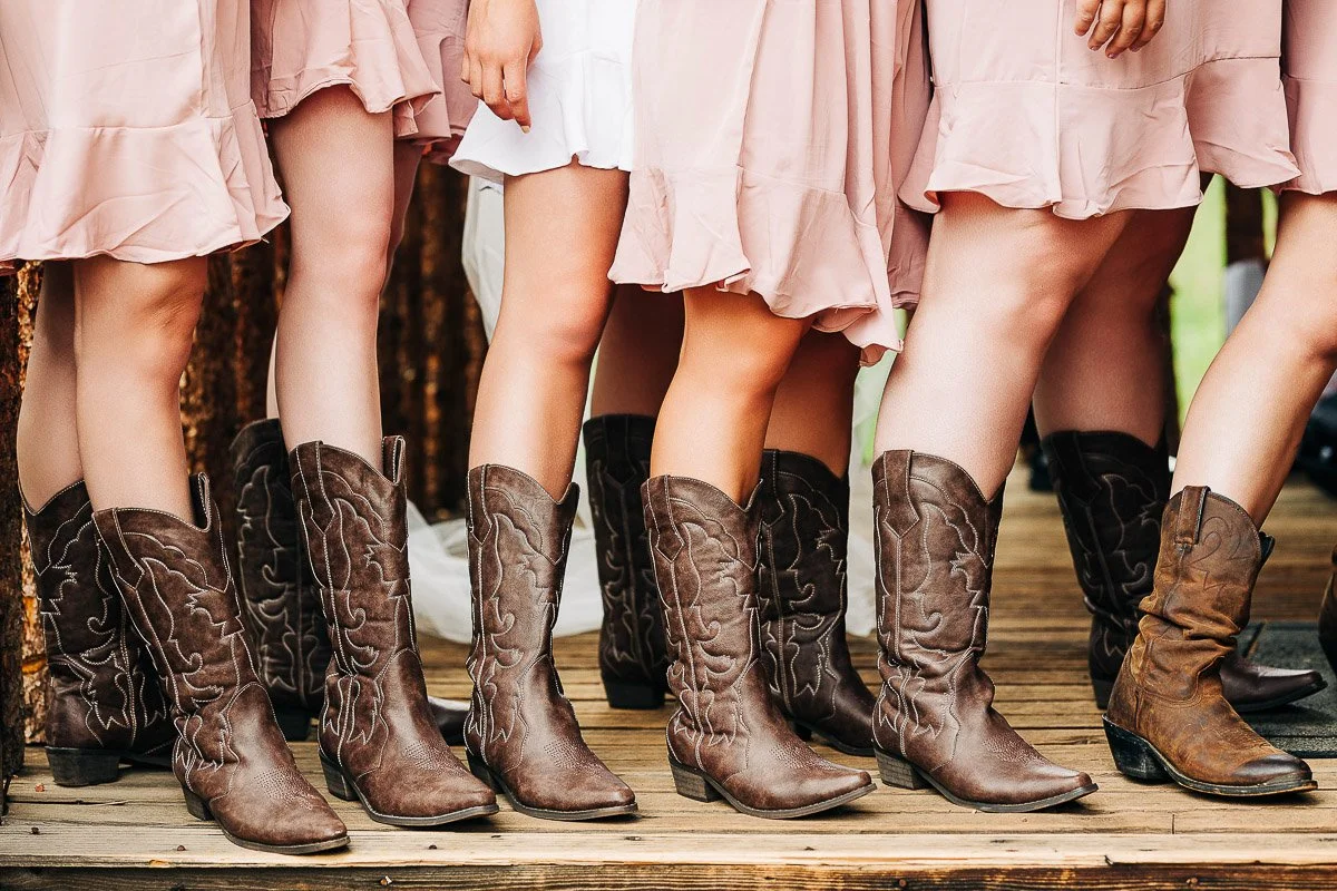 Six women in pink dresses and brown cowboy boots stand on a wooden deck. The scene conveys a rustic, coordinated, and casual atmosphere.