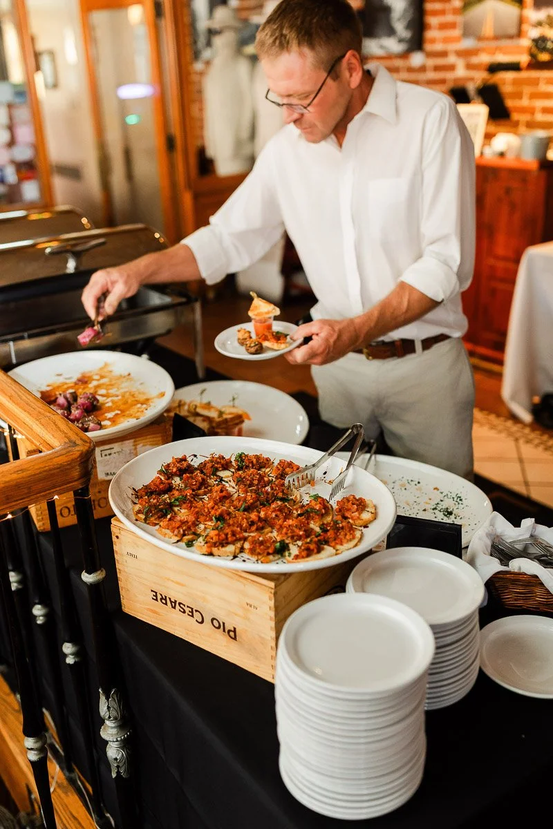 A man in a white shirt serves himself food from a buffet table. The setting is warm and inviting, with plates of colorful dishes and stacked plates.