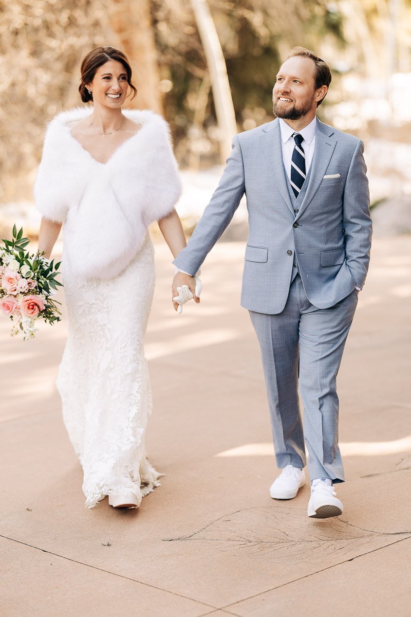 Smiling bride in a white gown and faux fur stole holds hands with groom in a light blue suit and sneakers, walking outdoors with a bouquet.