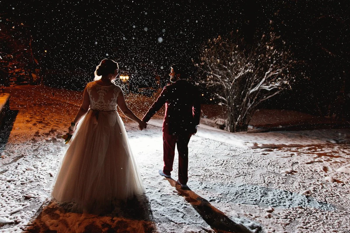 A couple holds hands at night, surrounded by softly falling snow. The bride wears a lace gown, the groom is in a dark suit. A serene, magical scene.