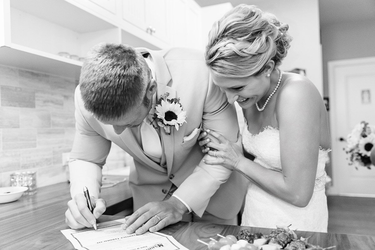 A bride and groom in a joyful moment; the groom is signing a document while the bride leans in, smiling warmly. Both in wedding attire, conveying happiness.