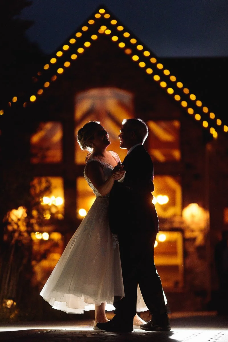 A bride and groom dance under soft lights, silhouetted against a warmly lit Della Terra Wedgewood with glowing windows. The scene is intimate and romantic captured by Estes Park Wedding Photographer tomKphoto