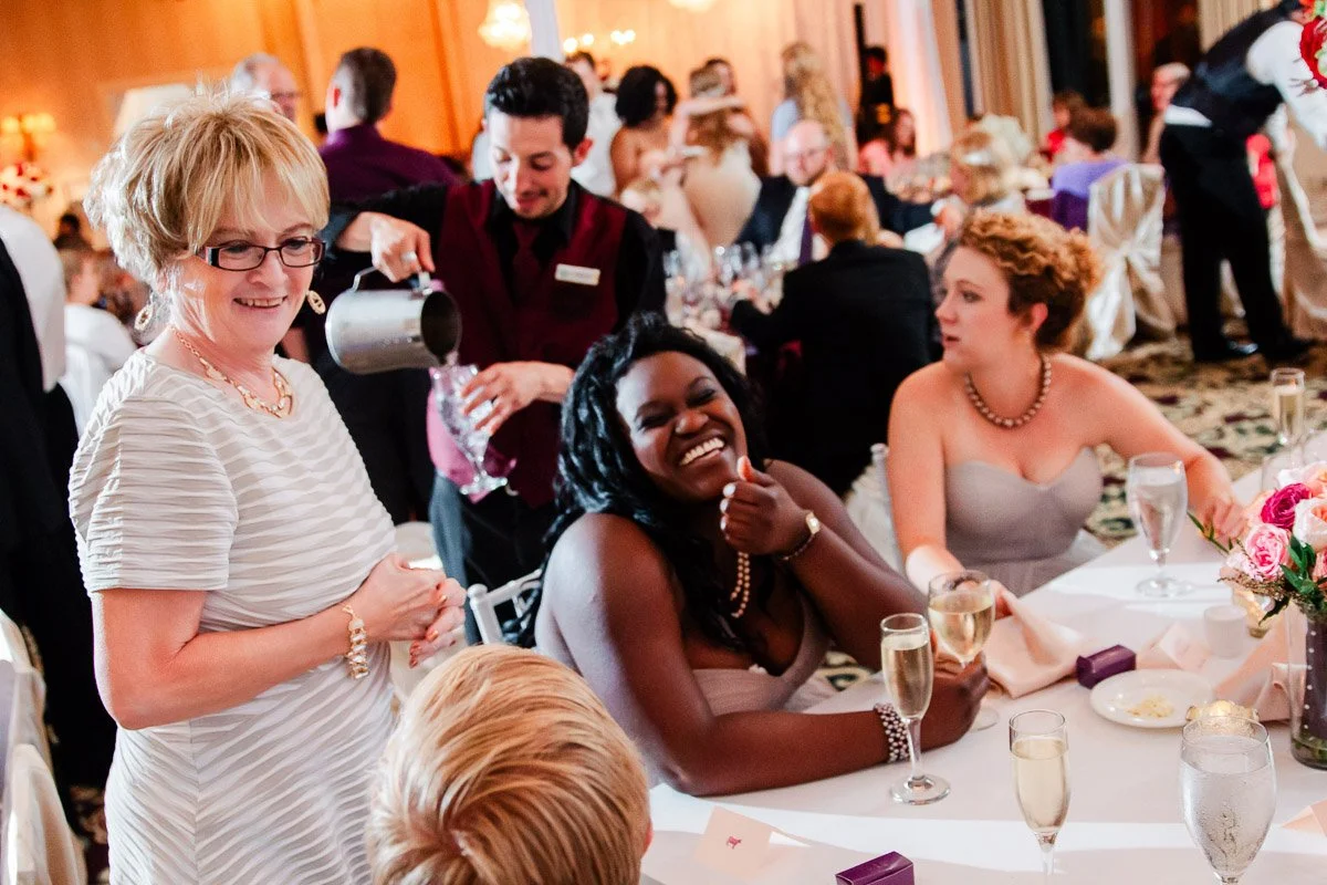 Guests at a wedding reception smile and chat around a table. A waiter pours water while guests enjoy champagne and conversation in a lively atmosphere.