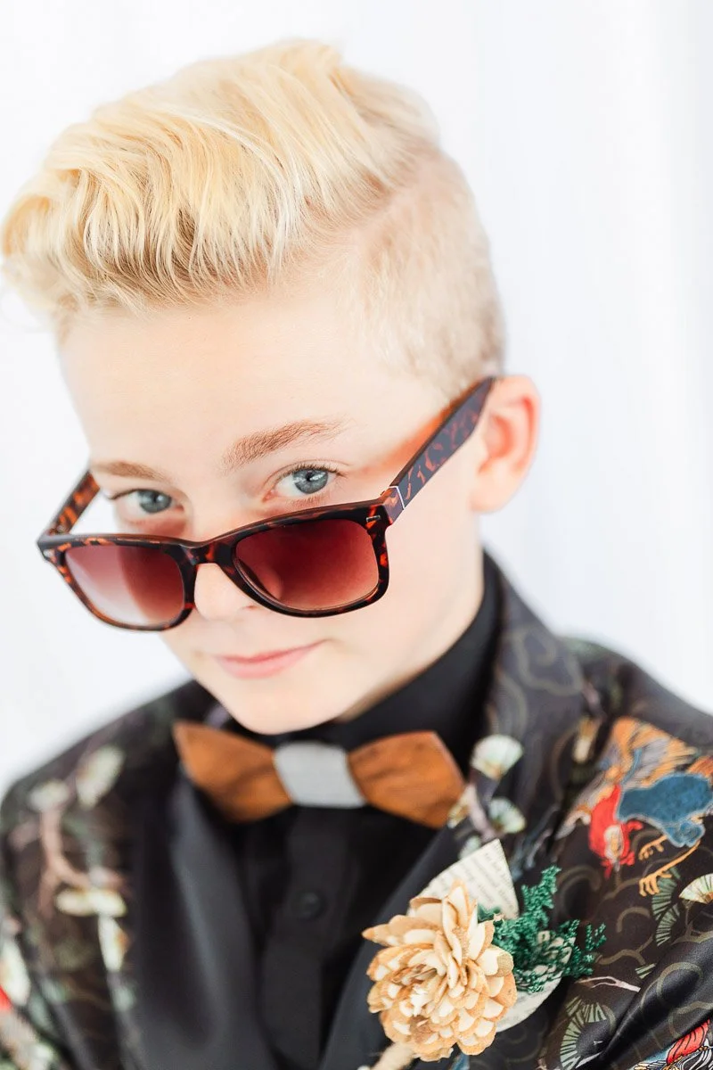 A confident young person with stylish blond hair wears large sunglasses and a floral-patterned suit. They have a playful expression, complete with a wooden bow tie and a pinecone boutonniere.
