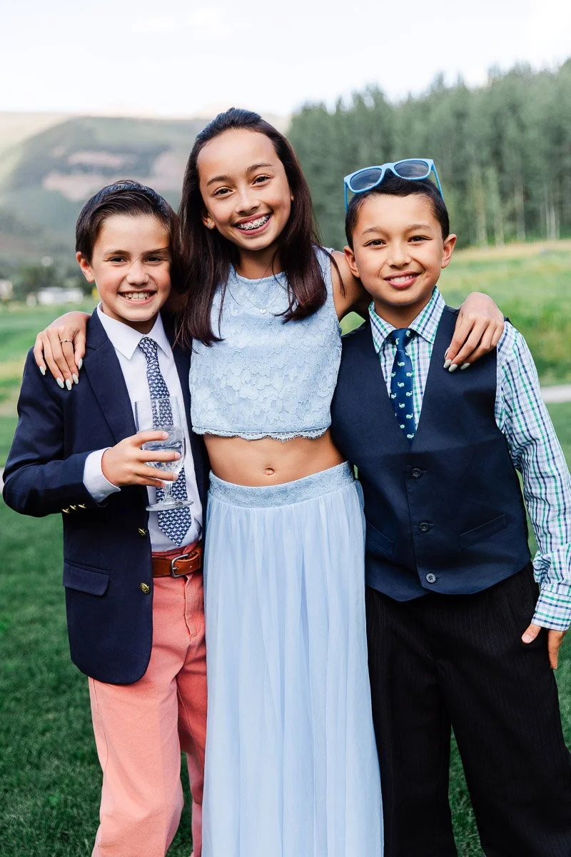 Three smiling children pose together outdoors. The girl in the middle wears a light blue dress, flanked by two boys in suits. A joyful, festive scene.