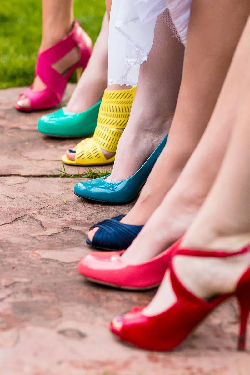 A row of colorful high heels on women’s feet lined up on a stone path. Shoes are red, blue, yellow, pink, and teal, conveying a lively, vibrant mood.