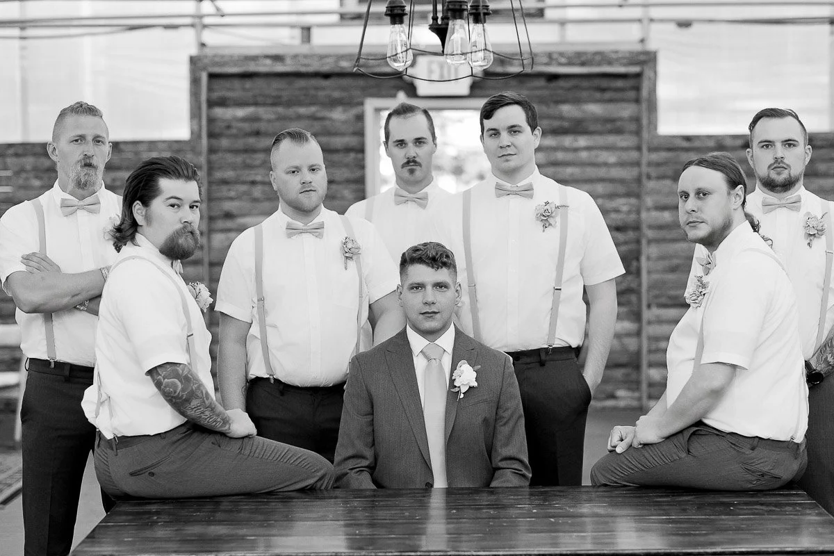 Eight men pose for a formal wedding photo. The groom in a suit sits while the groomsmen in white shirts with bow ties and suspenders stand and sit around him.