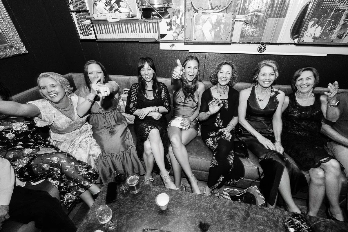 A group of seven women sit closely on a sofa, smiling and laughing. Some give thumbs up, creating a lively, celebratory atmosphere in a cozy room.
