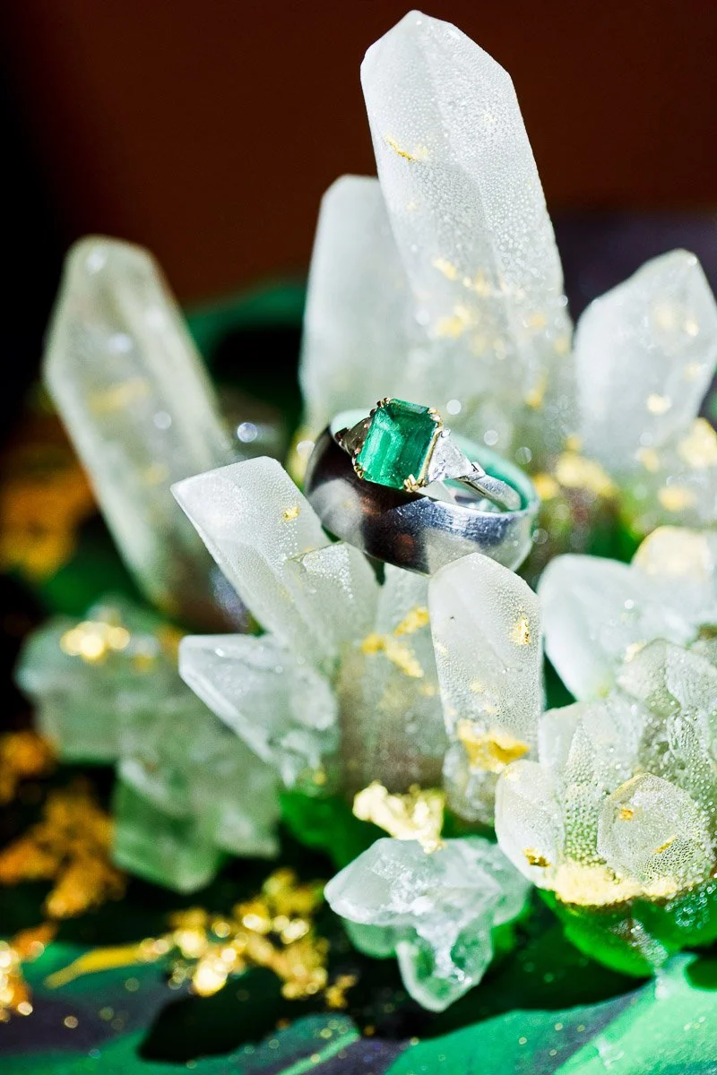 A vibrant emerald ring is elegantly placed on a cluster of clear crystal formations, dusted with gold flecks, creating a luxurious and enchanting scene.