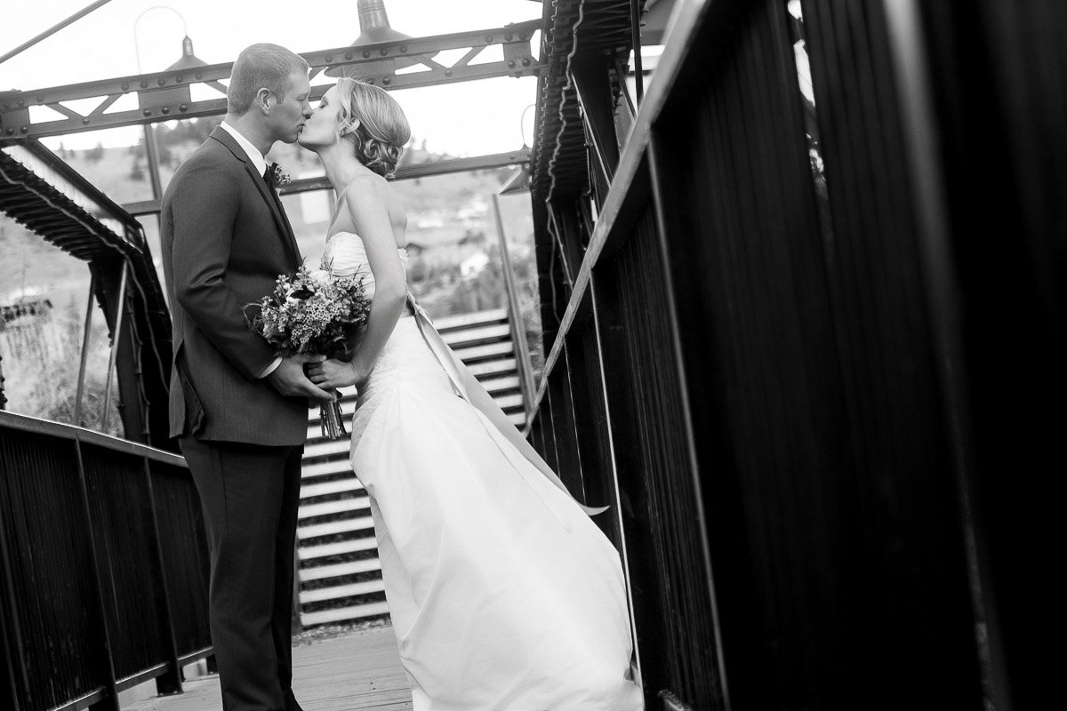 Black and white photo of a bride in a flowing gown and groom in a suit kissing on a metal bridge, conveying romance and timeless elegance.