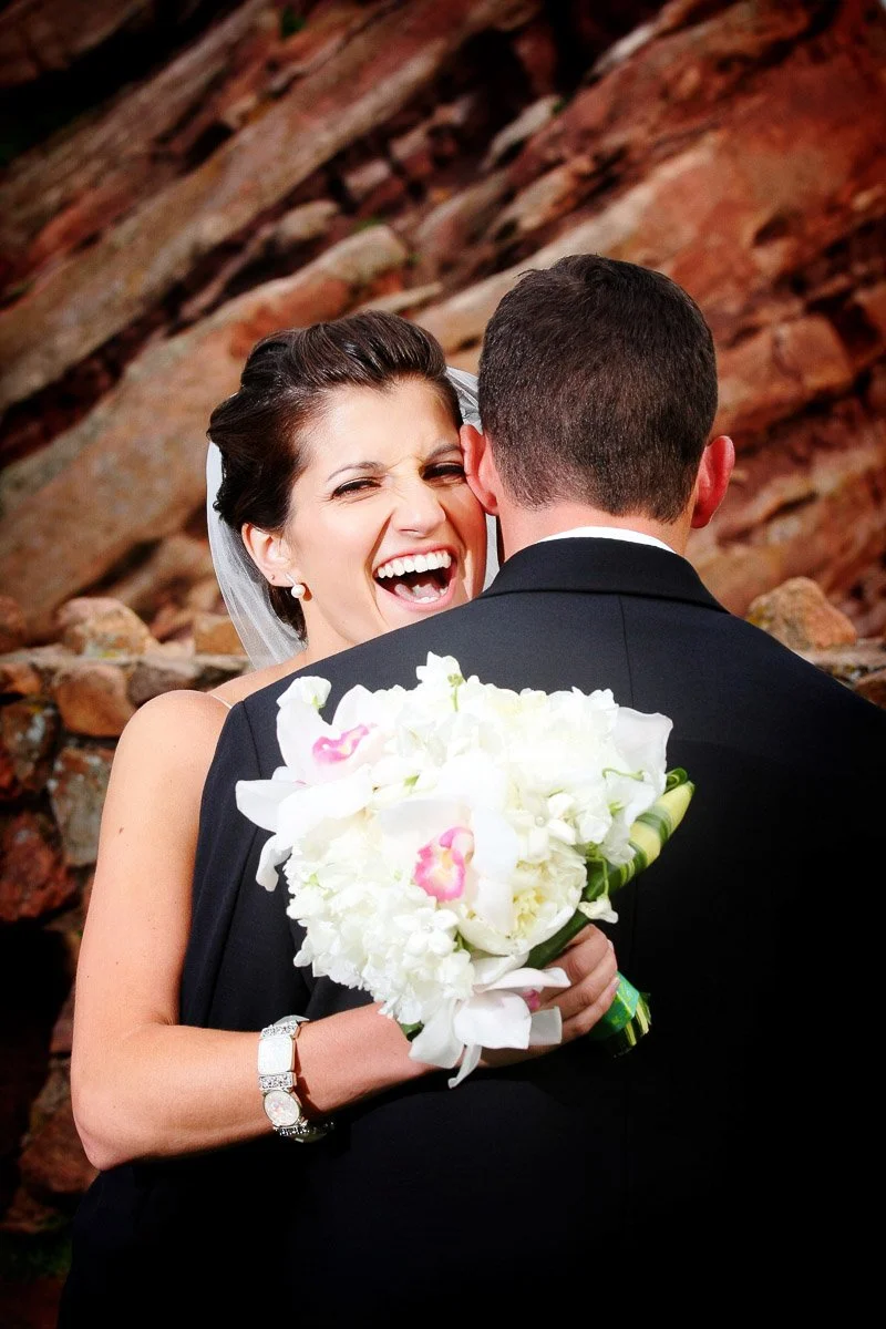 A bride joyfully laughs while hugging a groom in front of a rocky background. She holds a bouquet of white and pink flowers, conveying happiness.