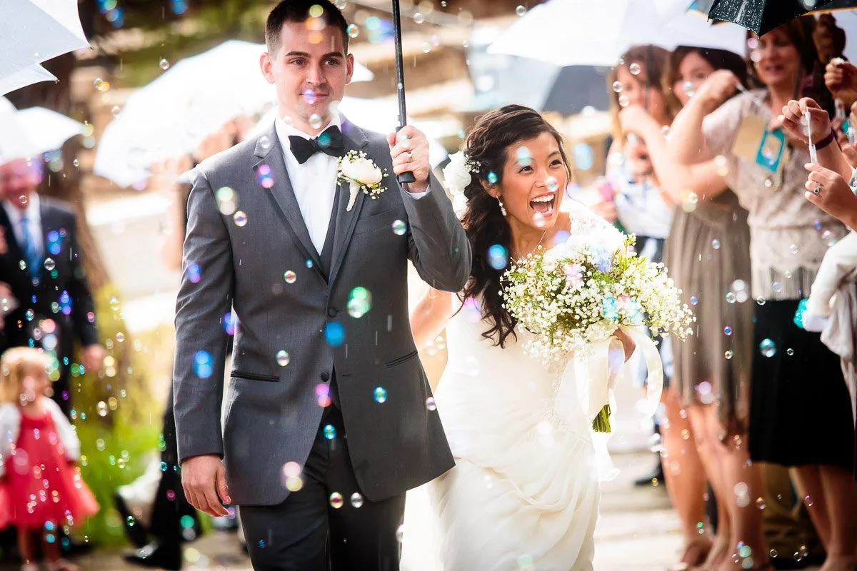 A joyous bride and groom walk under umbrellas through a shower of bubbles, surrounded by smiling guests. The scene is vibrant and celebratory.