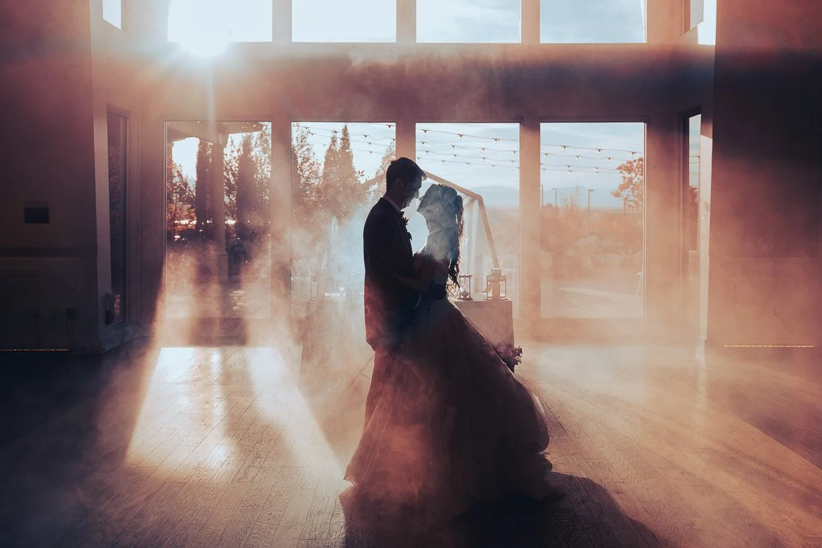 A bride and groom share a romantic dance in a sunlit room, surrounded by soft mist. Large windows reveal a scenic view, casting a warm, ethereal glow.