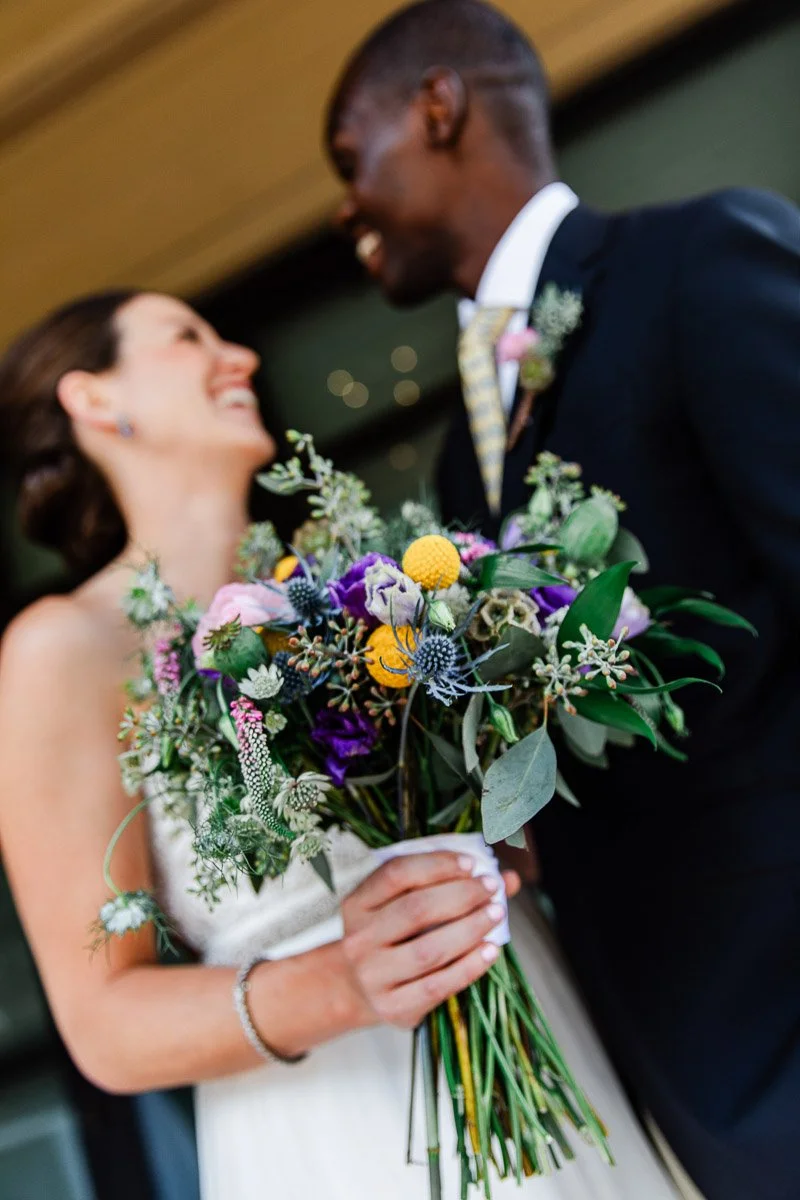 Bride and groom smiling at each other, with the bride holding a vibrant bouquet of yellow, purple, and pink flowers. The mood is joyful and intimate.