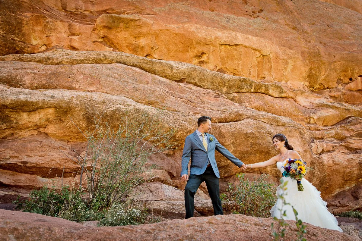 A bride and groom hold hands on rocky terrain with red sandstone cliffs in the background. The bride wears a white gown and holds a colorful bouquet.