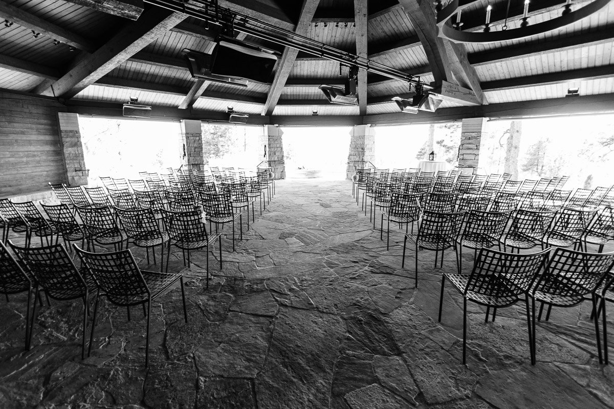 A black and white image of an empty, rustic auditorium with stone floors, lined with metal chairs facing a bright, open window. The mood is serene and contemplative.
