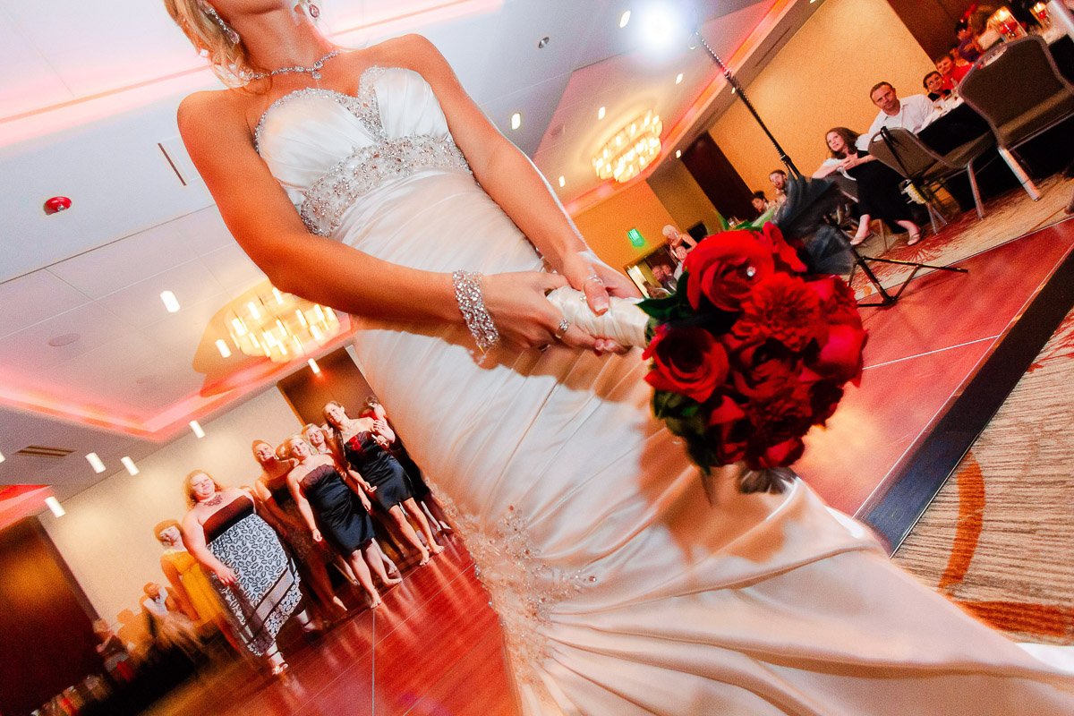Bride in elegant white gown holding red bouquet on a tilted dance floor, guests in background watching, ornate chandeliers add warmth.