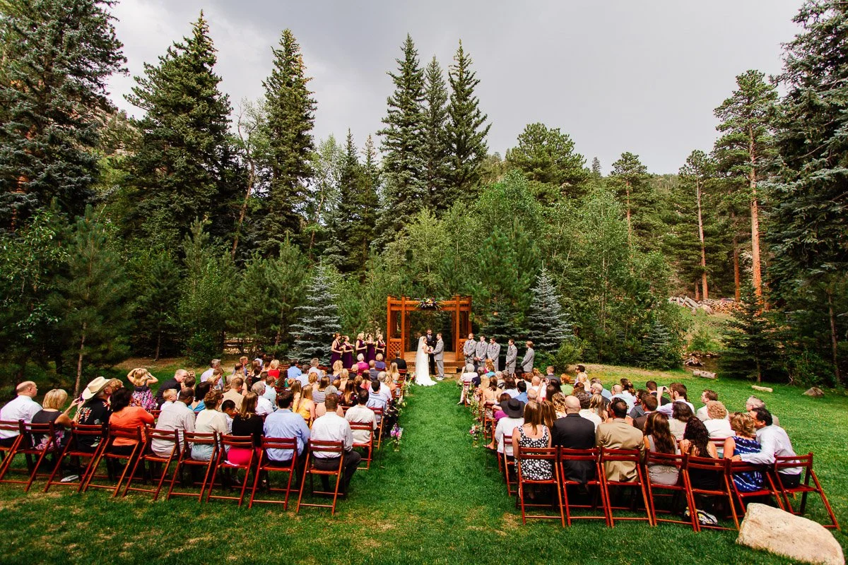 Outdoor Estes Park Condos wedding ceremony in a forest setting with tall evergreens. Guests sit on wooden chairs facing a couple under a wooden arch. The mood is serene and joyful.