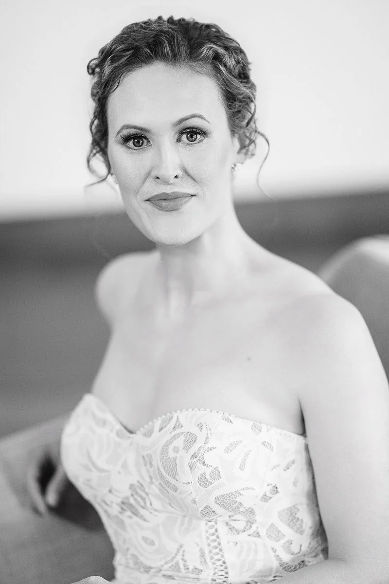 Black and white portrait of a woman in an elegant lace dress. She sits gracefully, gazing softly at the camera with a subtle smile and poised demeanor.
