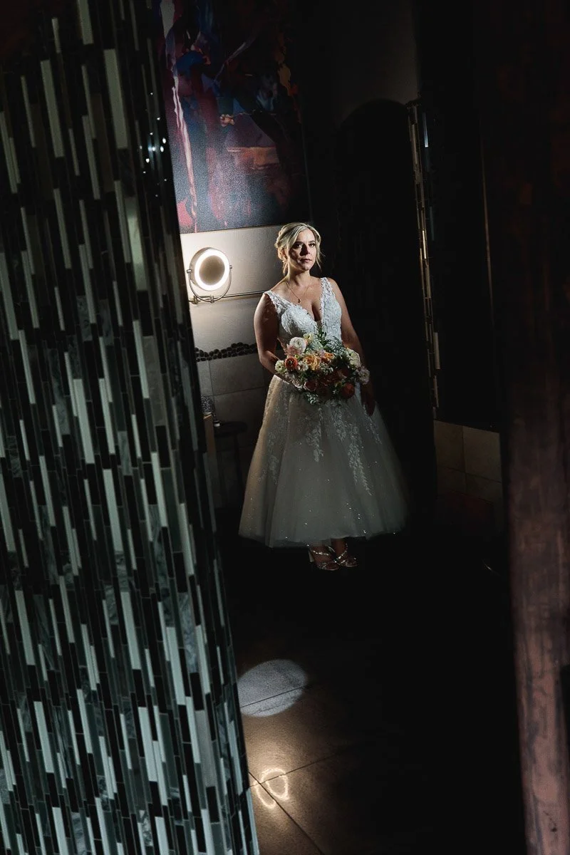 A bride in a white lace gown stands holding a bouquet of flowers during a Della Terra wedding. Soft lighting casts a warm glow, creating an intimate and serene atmosphere.