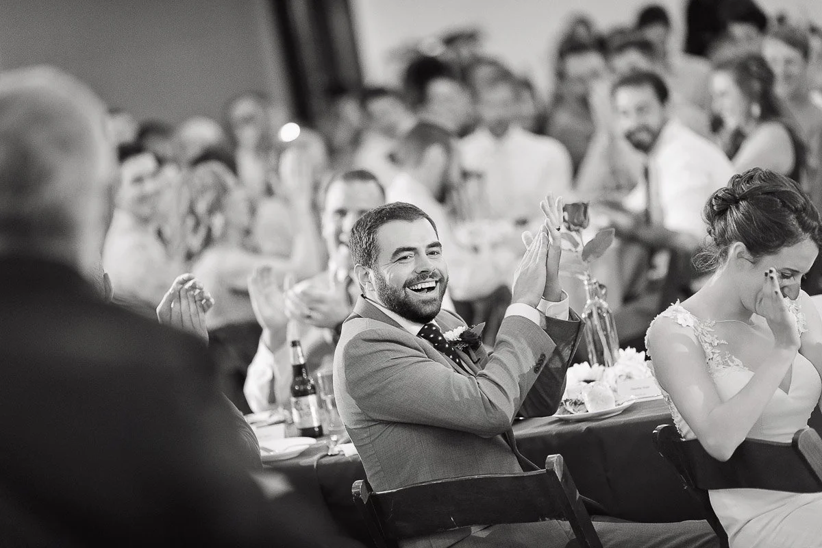 A joyous wedding scene with a man in a suit laughing and clapping at a crowded table. Guests in the background applaud, creating a celebratory atmosphere.