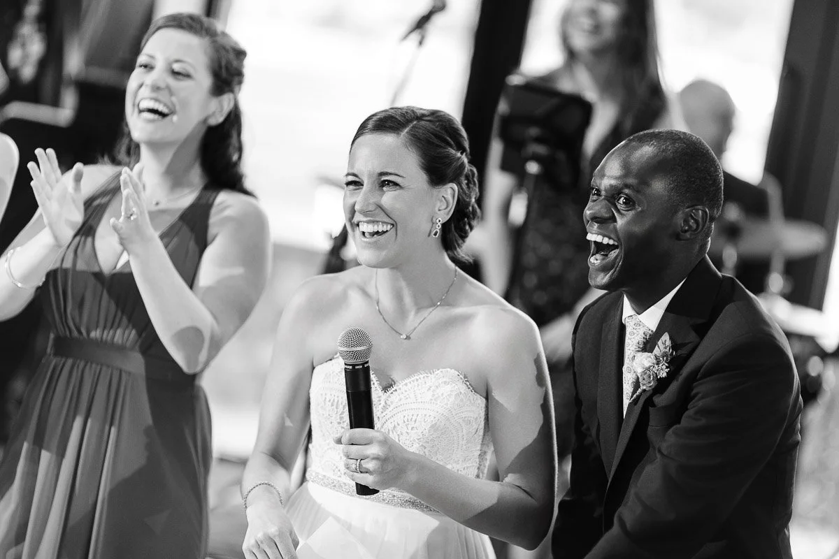 A joyful bride in a white dress, holding a microphone, stands between a laughing groom in a suit and a clapping bridesmaid. Black and white photo.