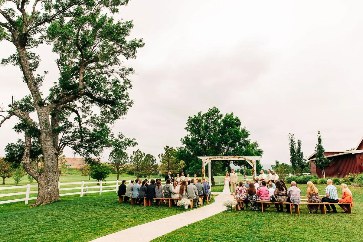 A serene outdoor wedding ceremony on a lush green lawn with guests seated on wooden benches. An elegant archway stands in front, framed by trees.