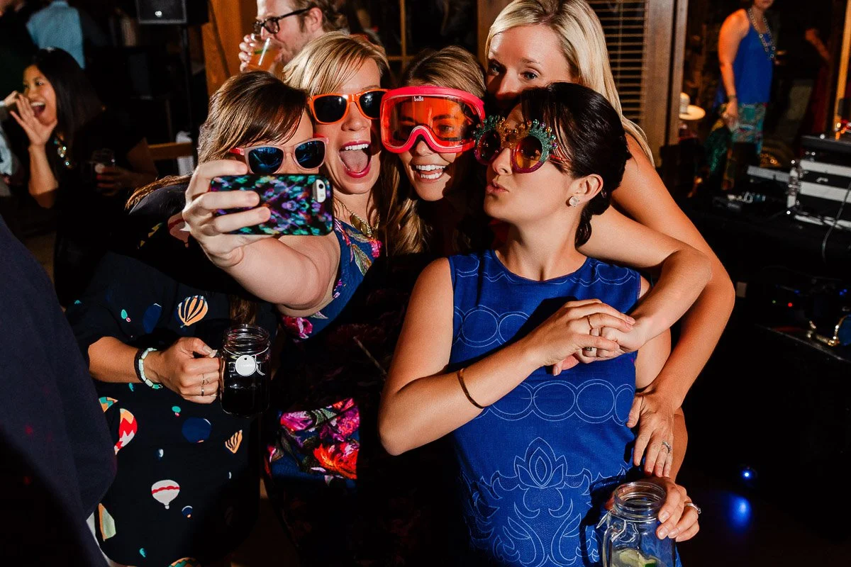 Five women, dressed in colorful party attire and novelty glasses, joyfully pose for a group selfie at a lively indoor party.